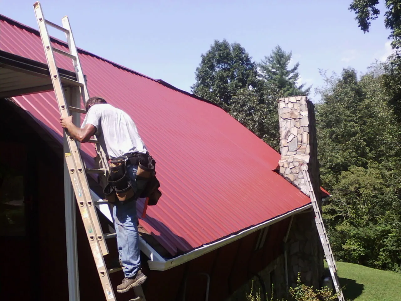 Roofer on a ladder beside a red metal roof and stone chimney — Soffit & Fascia Repair by Roofing Brothers in Maurice